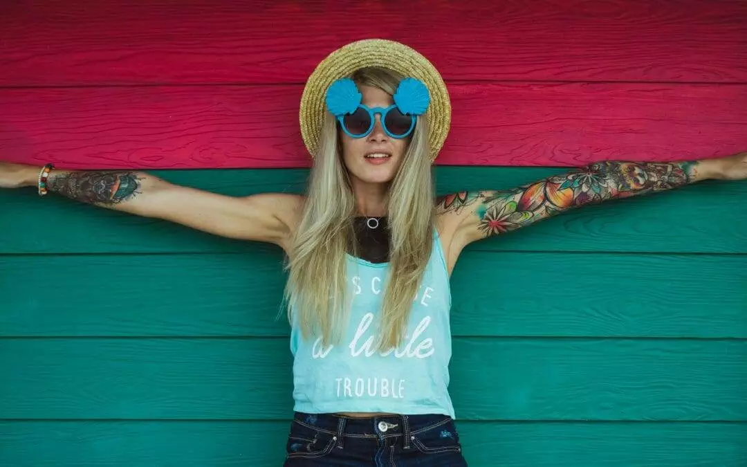 A blonde woman with tattoos, wearing a straw hat, round blue sunglasses, and a light blue tank top, stands against a red and green background with her arms outstretched.