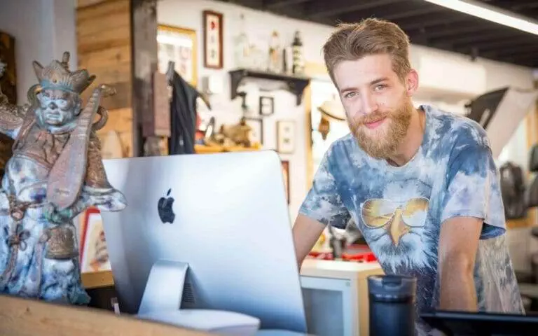 A man with a beard wearing a t-shirt featuring an eagle stands behind an Apple computer in a room decorated with various artifacts.