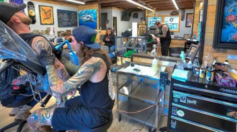 Several people are getting tattooed in a tattoo studio. Two tattoo artists are working on clients, while others are interacting in the background. The studio is equipped with various tattoo supplies and decorations.