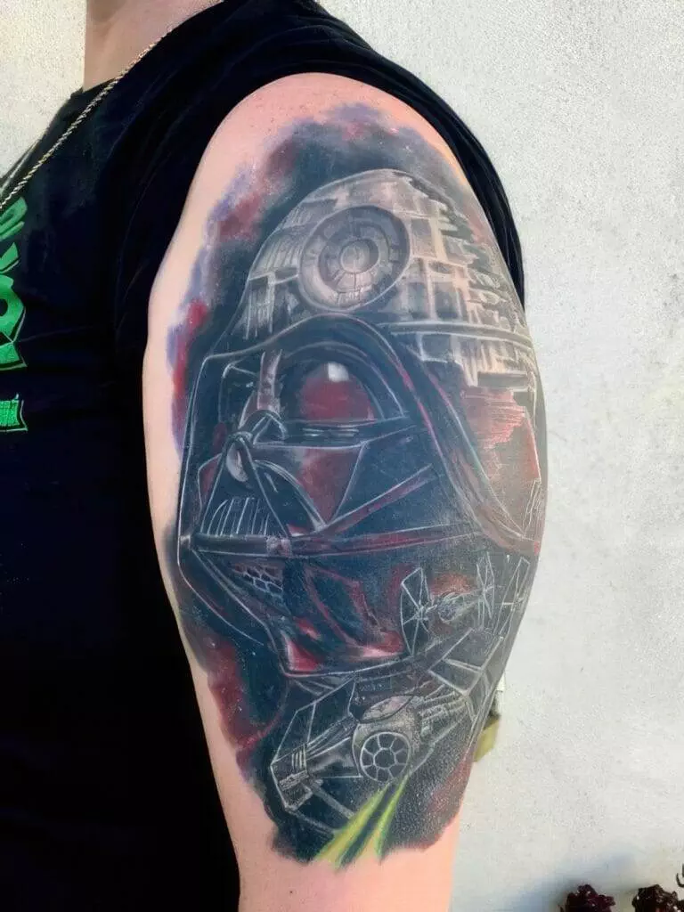 Close-up of a person's upper arm featuring a detailed tattoo of Darth Vader, TIE fighters, and the Death Star from Star Wars. The person is wearing a black sleeveless shirt.