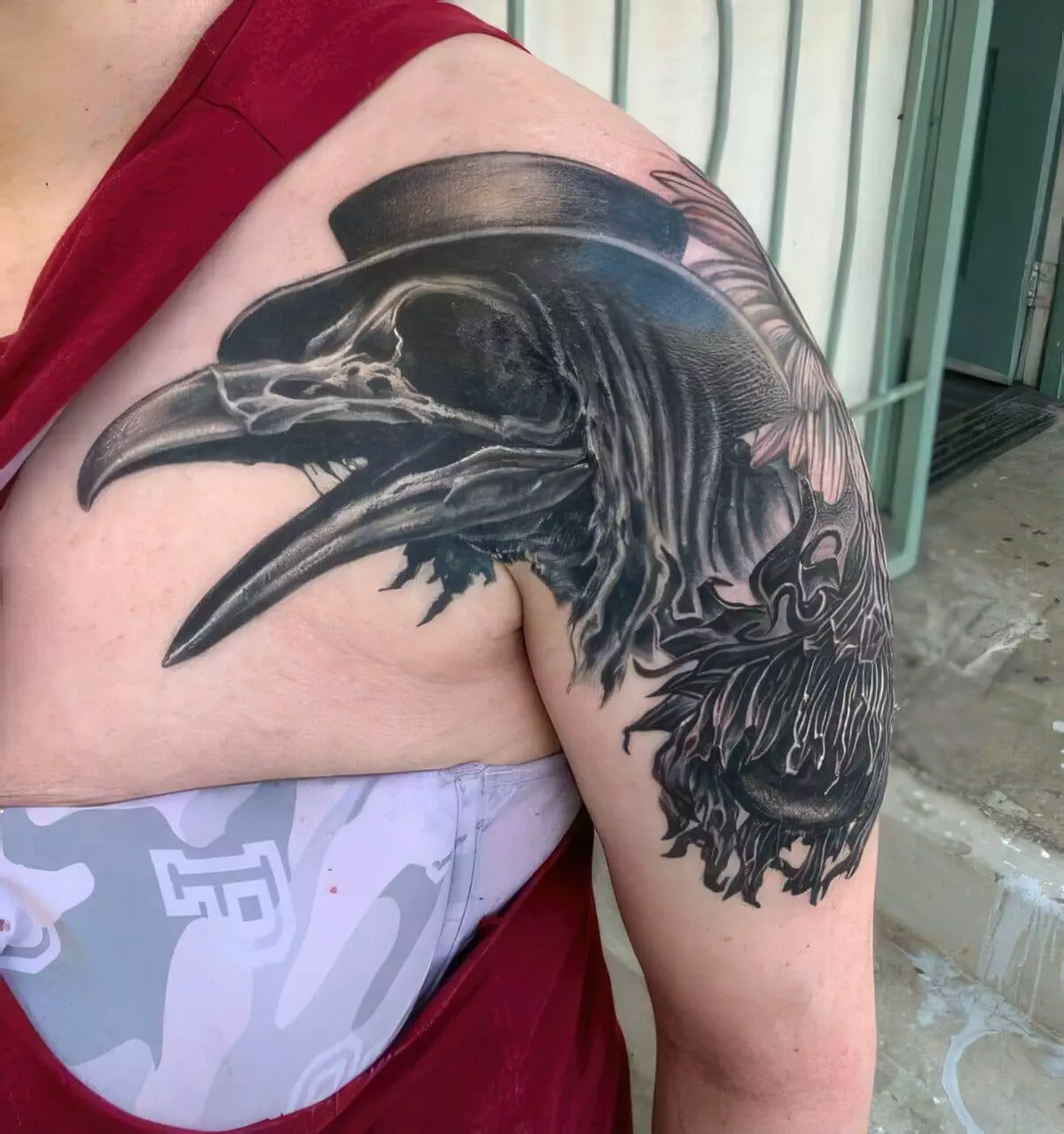 A person's upper arm and shoulder displaying a large black-and-grey tattoo of a raven wearing a top hat. The person is wearing a red sleeveless shirt.