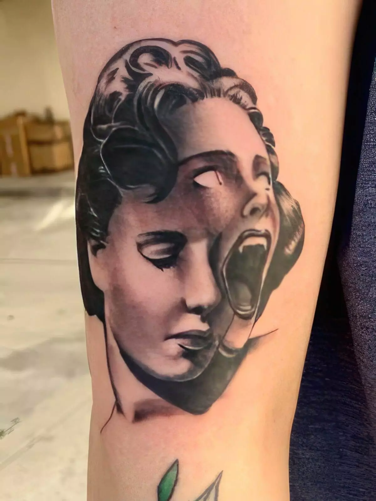 A tattoo of a person's face, with one side calm and the other side screaming, is displayed on a person's arm. The design is in black and gray tones.