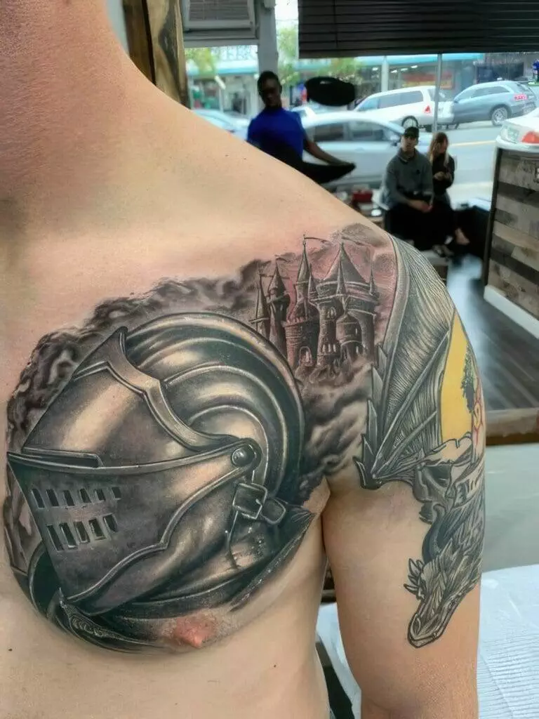 Close-up of a person's upper chest and shoulder showcasing a detailed tattoo of a knight's helmet, a castle in the background, and a dragon on their arm.