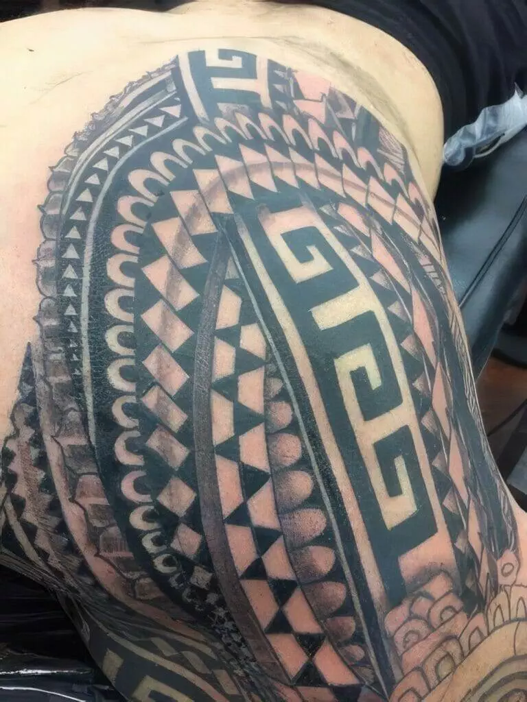 A close-up of an intricate black and grey tribal tattoo covering a person's side and part of their back. The design includes geometric patterns and symbols.