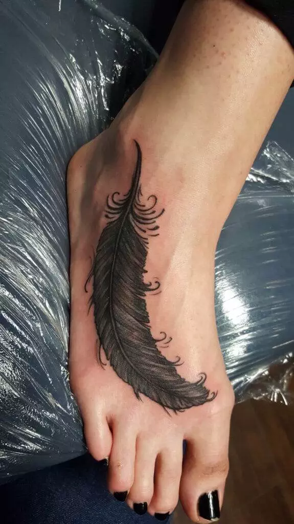 A tattoo of a black feather is seen on the top of a person's left foot, which is resting on a plastic-covered surface. The person's toenails are painted black.