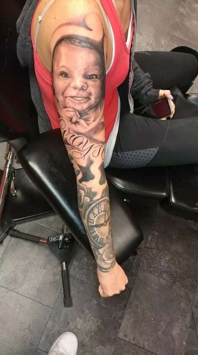 A person with a detailed tattoo of a baby's face and various designs covering the entire arm sits in a chair, wearing a red top and holding a smartphone.