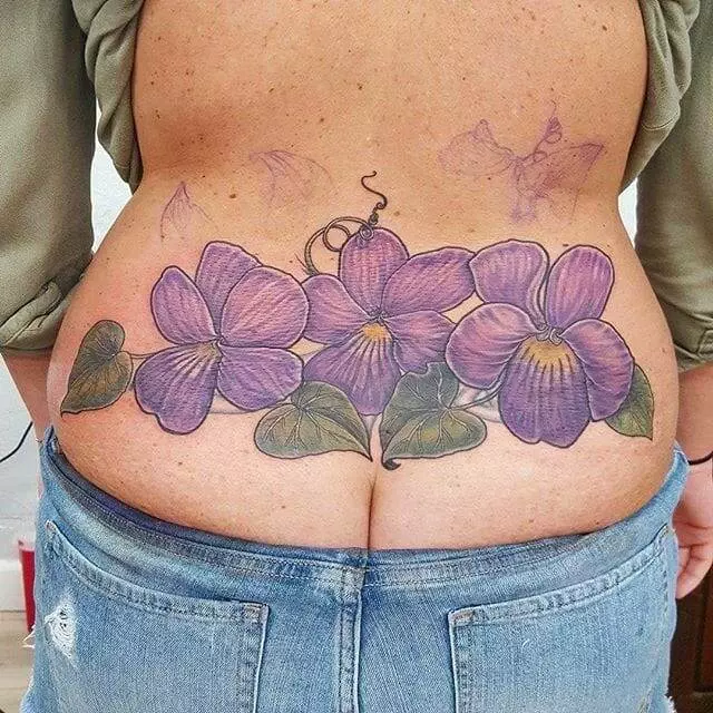 A lower back tattoo featuring three large purple flowers with green leaves and a butterfly on top, visible on a person wearing low-rise jeans and a partially lifted shirt.