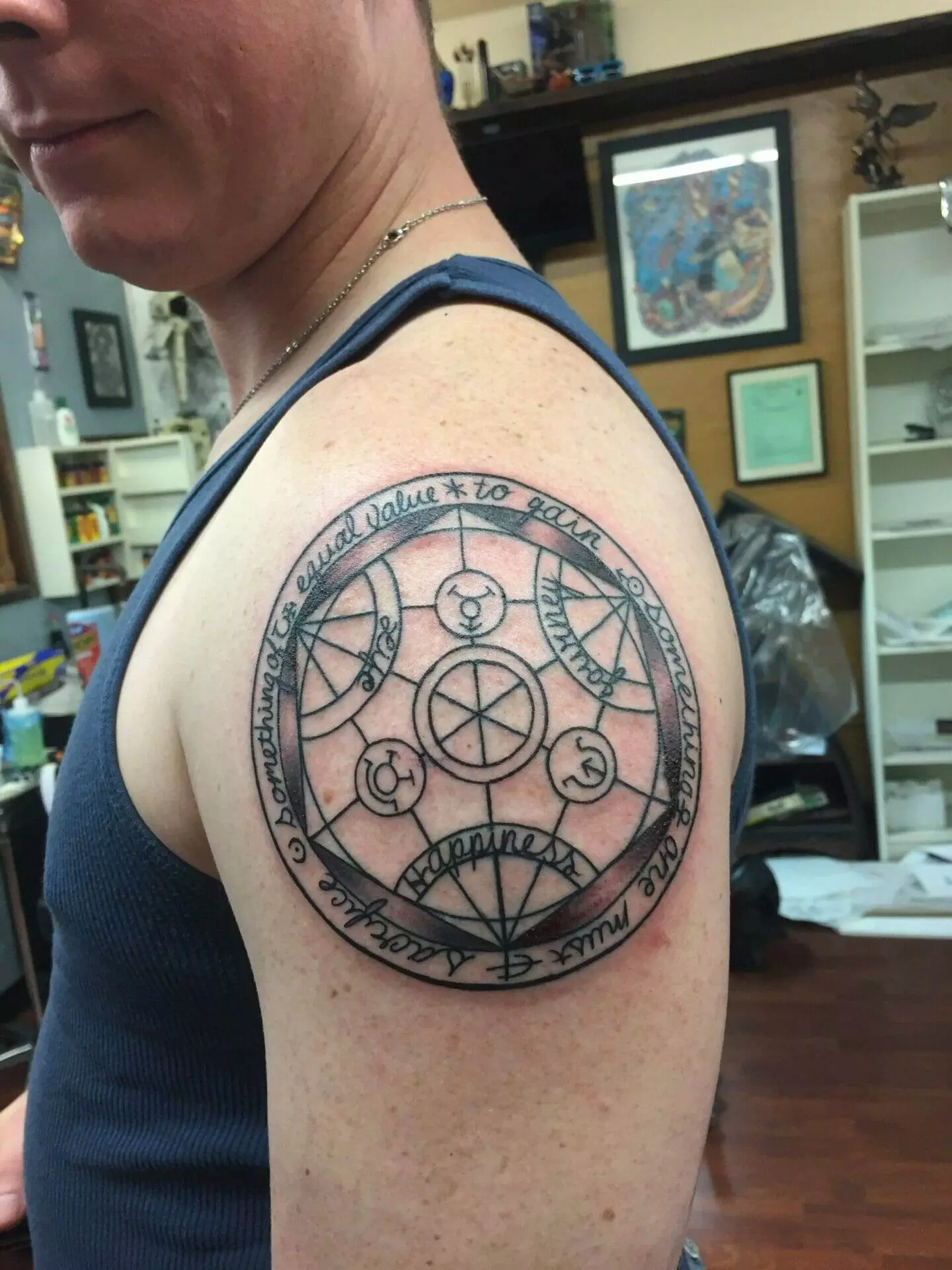 A person has a complex, geometric tattoo with circular designs and text on their upper arm. They are wearing a sleeveless shirt in an indoor setting.