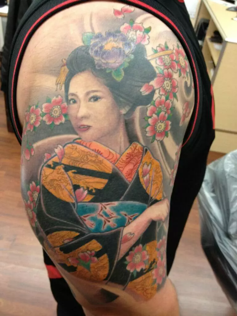 A detailed tattoo of a woman in traditional Japanese attire, featuring colorful flowers and intricate patterns, is seen on a person's shoulder and upper arm.