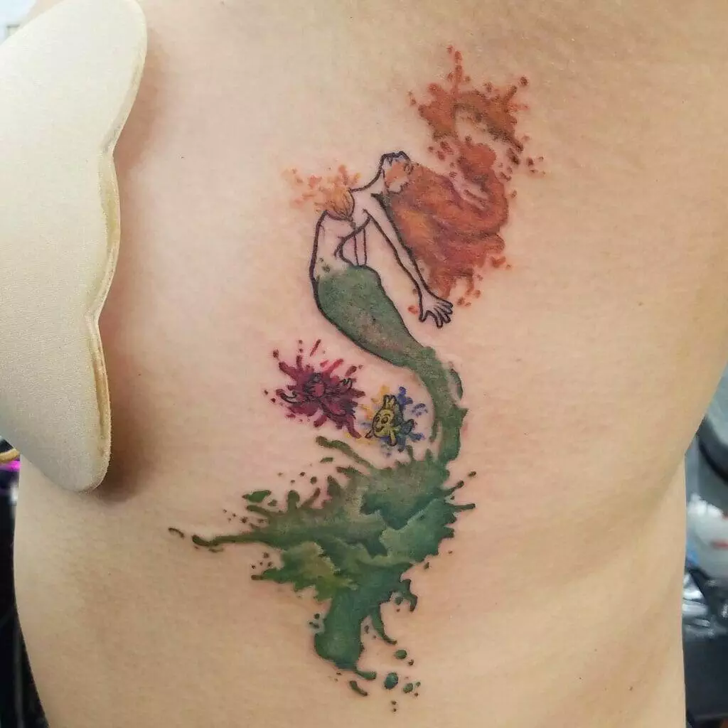 A colorful tattoo on skin depicts a mermaid with flowing red hair, flanked by two small sea creatures below her, one purple and one yellow.