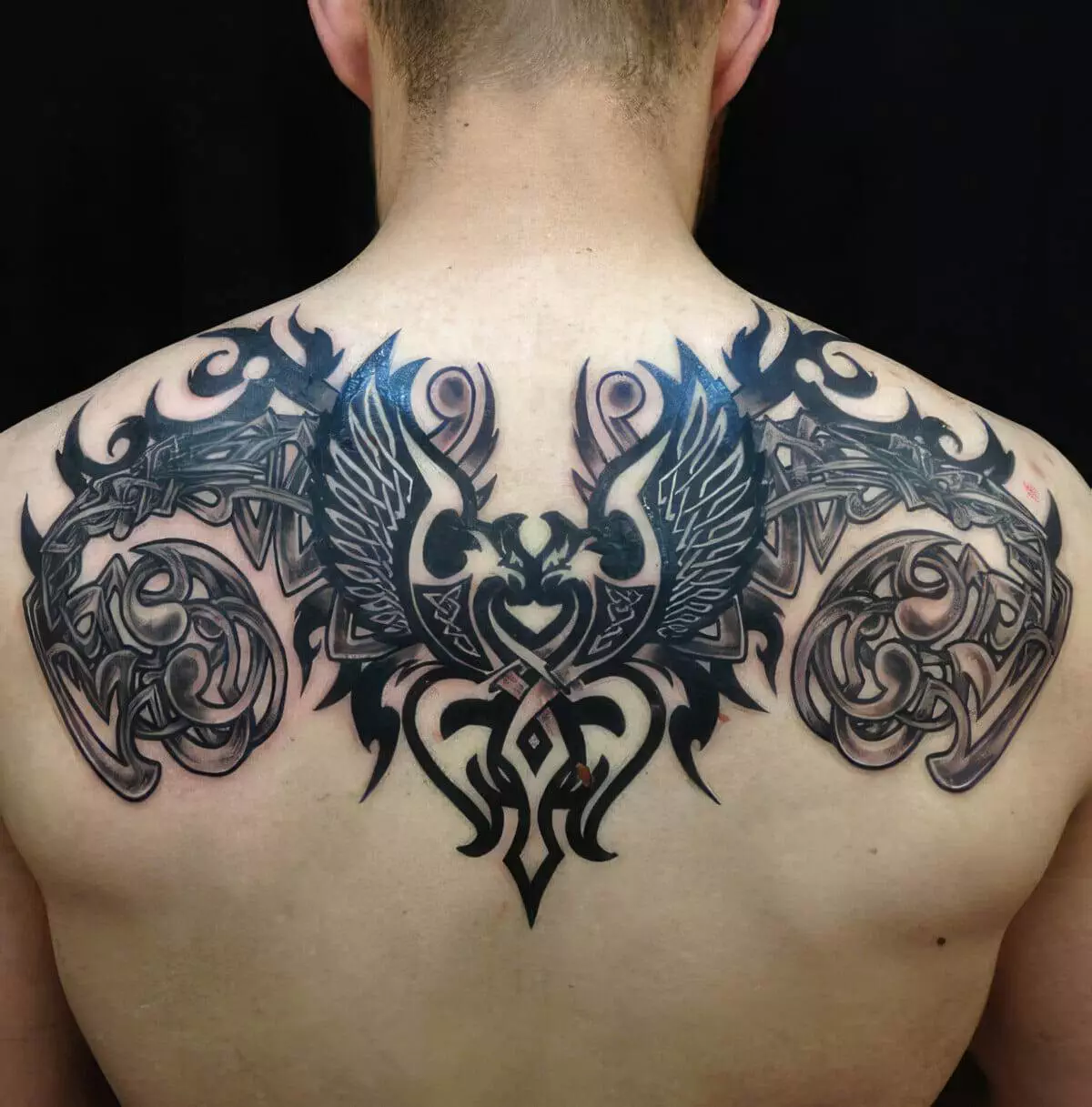 A person's back featuring an intricate black and dark blue tattoo design of symmetrical tribal patterns with wings in the center.