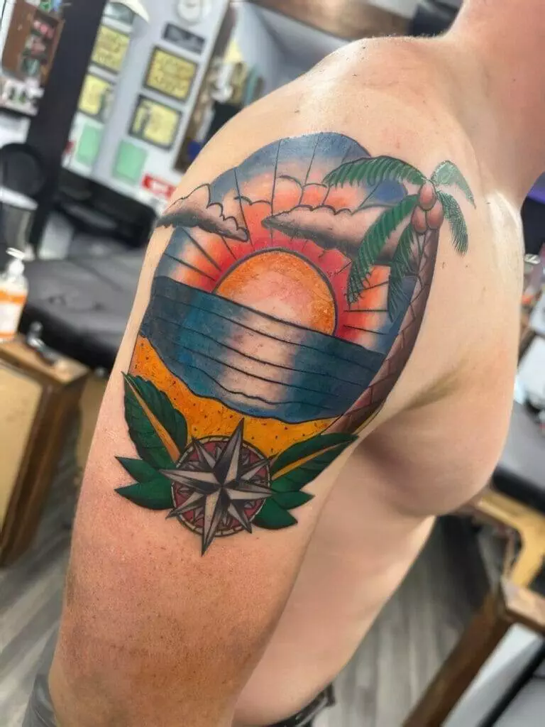 A colorful tattoo on an upper arm depicting a sunset over the ocean with palm trees, waves, and a compass rose at the bottom.