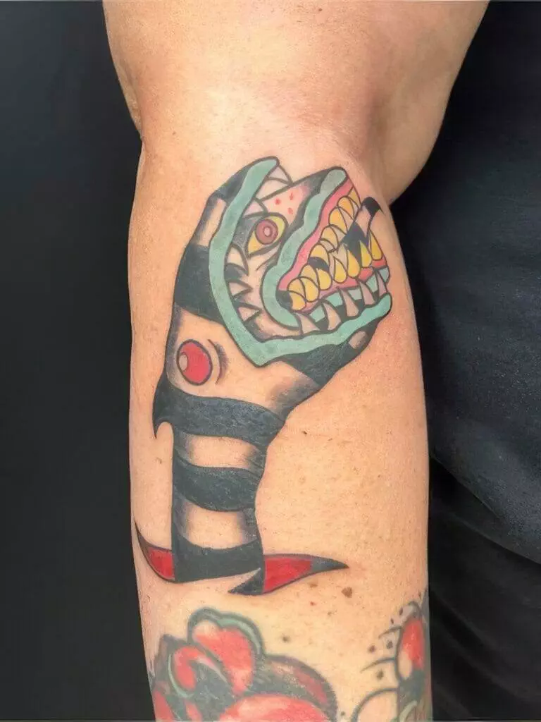 Colorful tattoo of a stylized snake with an open mouth showing sharp teeth, wrapped around and pointed downward on a person's arm, against a black background.