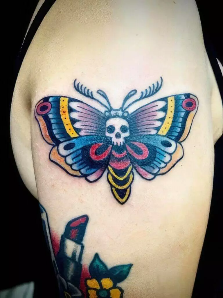 A colorful tattoo of a moth with a skull on its thorax is displayed on a person's upper arm.