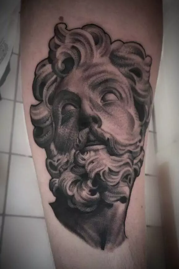 A tattoo of a grayscale ancient statue's head with curly hair and a beard on a person's arm.