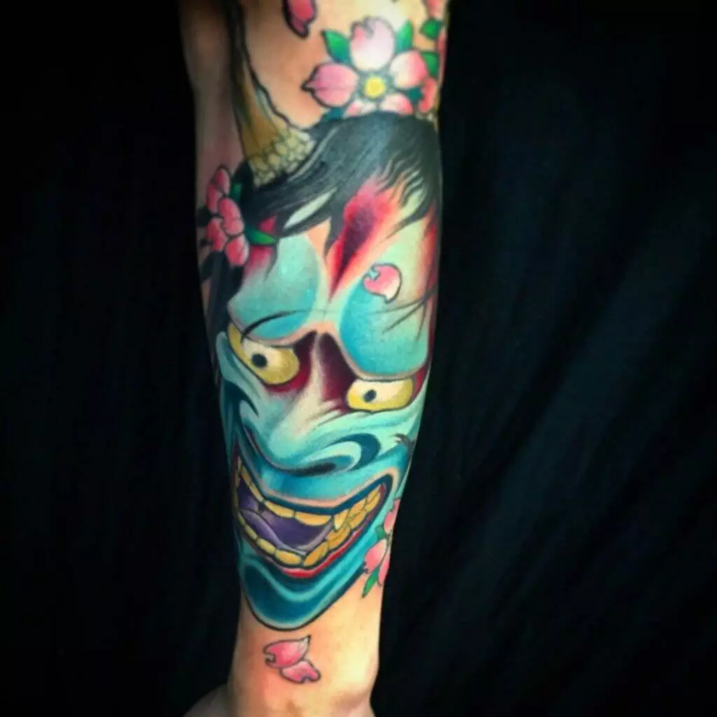 A colorful tattoo depicts a traditional Japanese Hannya mask surrounded by cherry blossoms on a person's forearm.