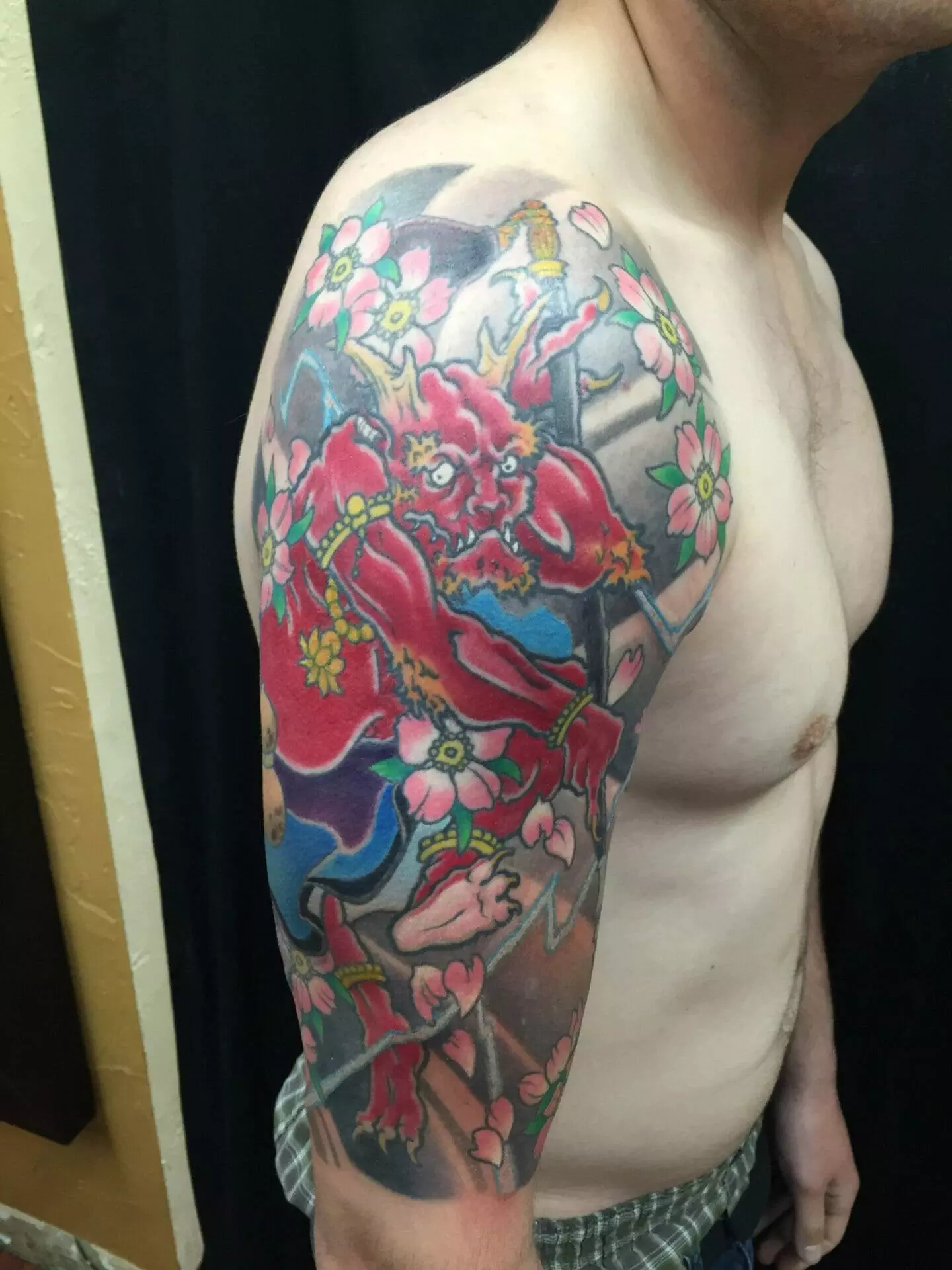 A person with a detailed and colorful tattoo of a red demon and flowers on their upper arm and shoulder stands against a dark background.