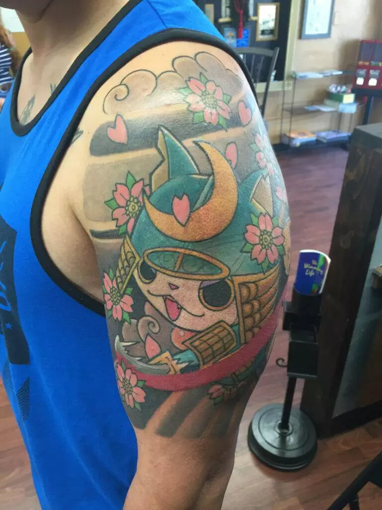 A colorful tattoo of a cartoonish samurai cat with cherry blossoms and hearts on the upper arm of a person wearing a blue sleeveless shirt.