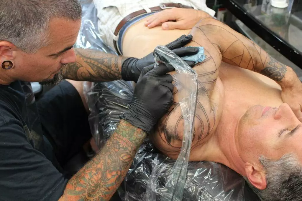 Tattoo artist with tattoos on arms and gauged ears, wearing black gloves, tattoos the upper chest and arm of a shirtless man lying on a table.