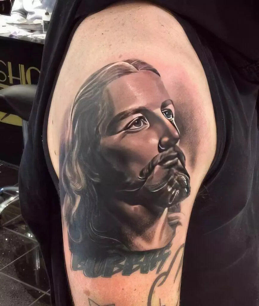 A detailed tattoo of a bearded man's face with long hair is displayed on a person's upper arm, showcasing realistic shading and meticulous design.