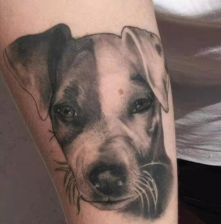 A realistic tattoo of a dog’s face, featuring detailed shading and highlights, is displayed on a person’s arm.