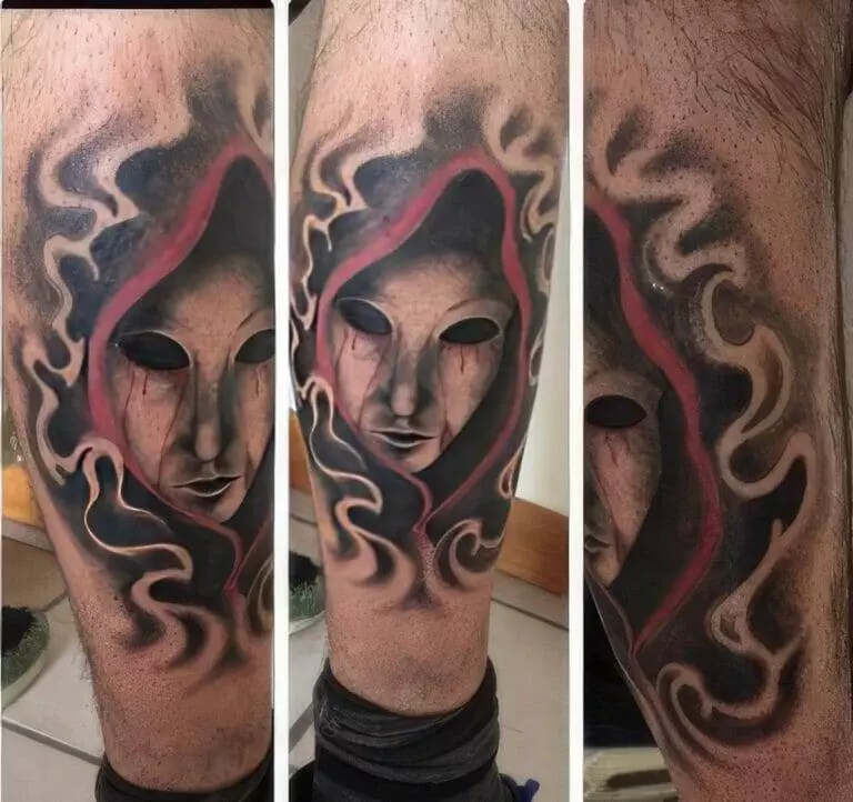 A tattoo of a hooded, mask-like face with dark eye sockets and red, tear-like marks, engulfed by swirling patterns.