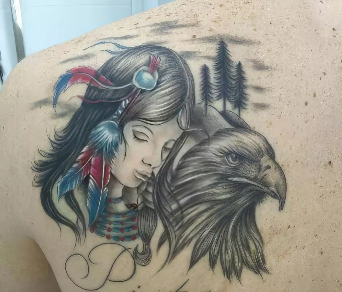 Tattoo on a person's upper back features a woman with closed eyes and feathered headband beside a detailed eagle's head with trees in the background.