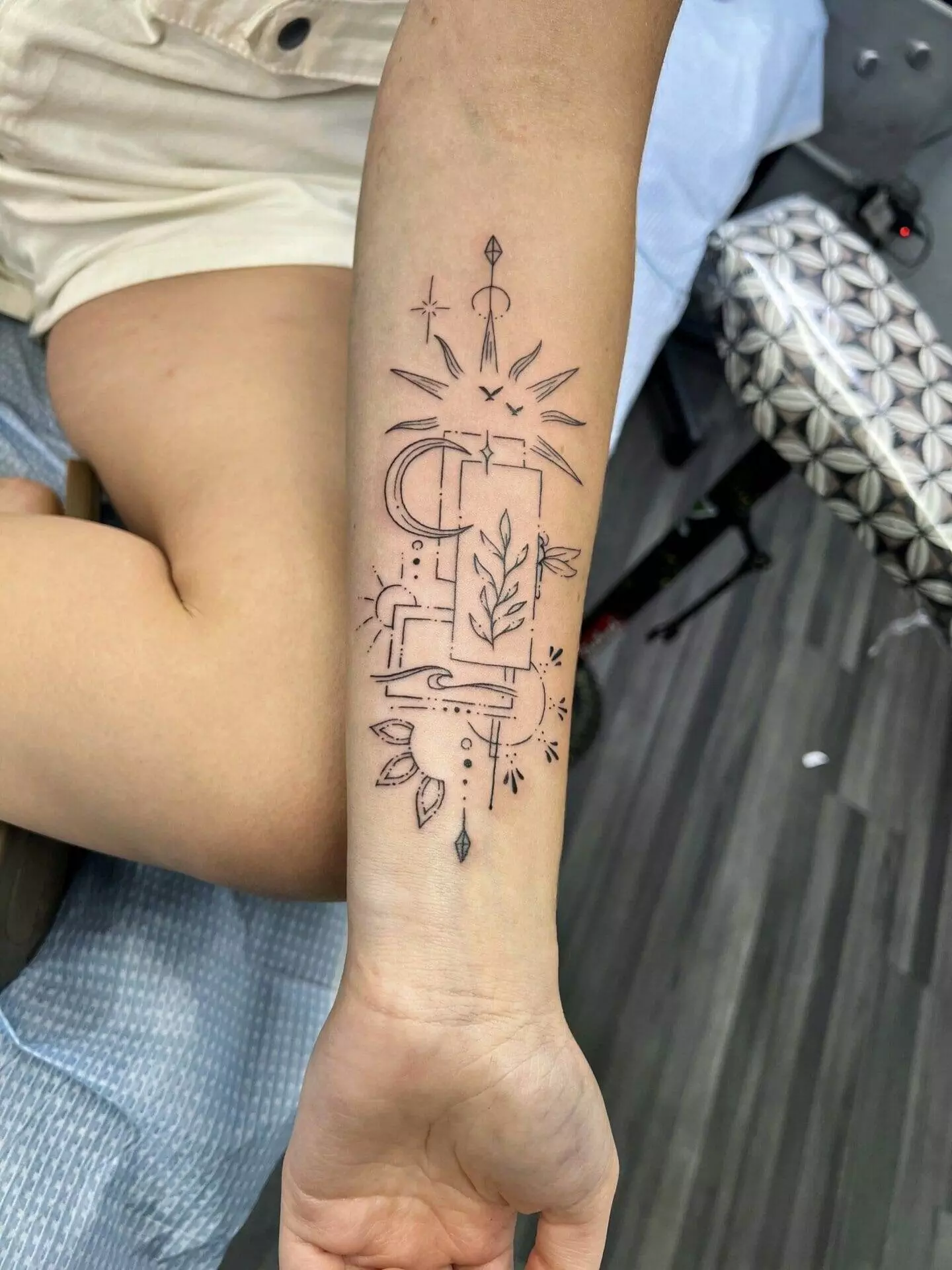 Forearm tattoo featuring abstract geometric shapes, botanical elements, a crescent moon, and small stars. The design is intricately detailed with a mix of lines and shading.