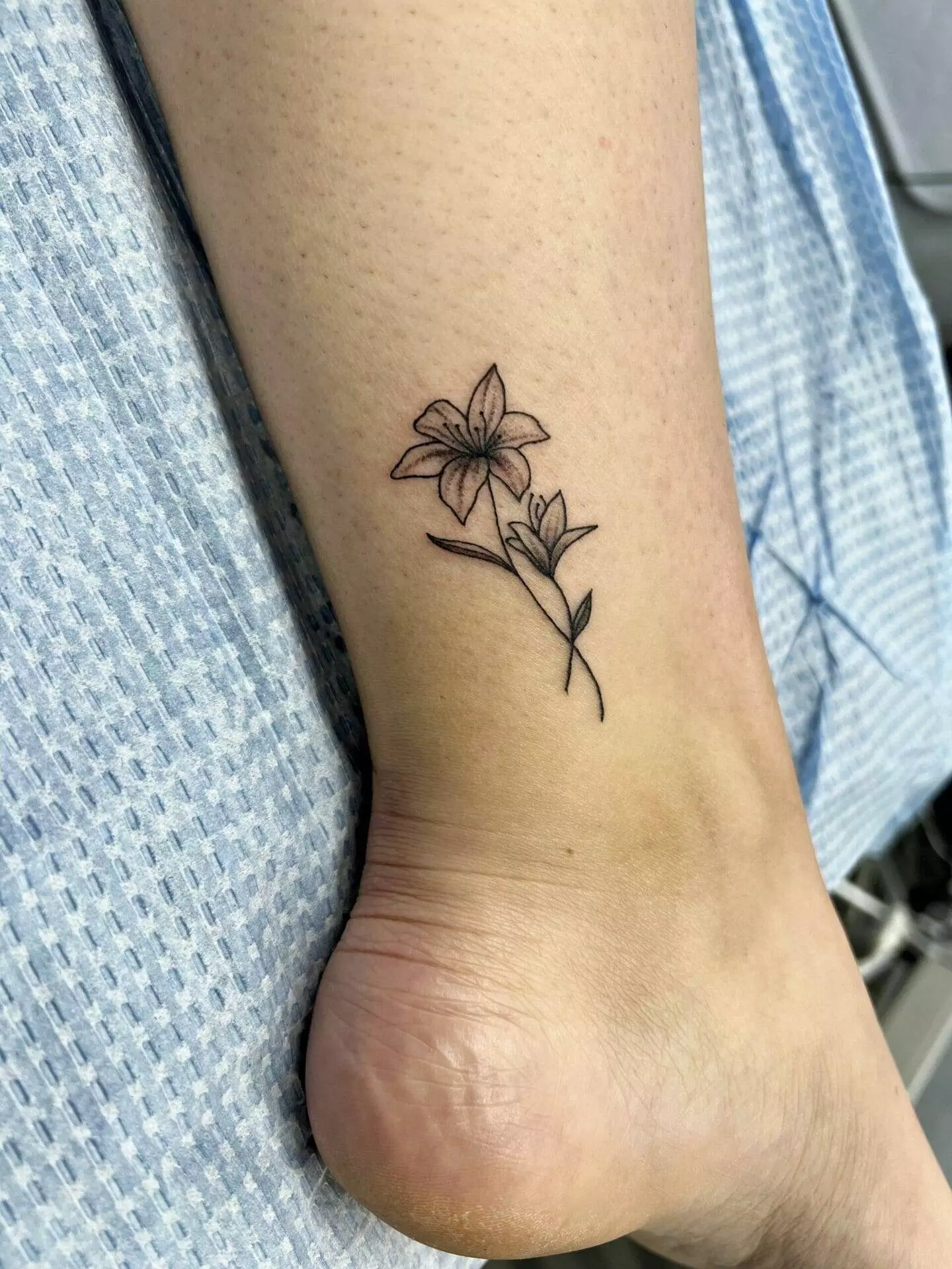 A delicate flower tattoo is visible on the inner ankle of a person. The design features a single flower with leaves, and the person is wearing a light blue, checkered garment.