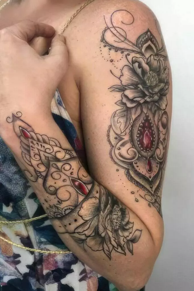 A close-up of an arm with intricate tattoos featuring floral designs and red gem elements, extending from the upper arm to the forearm. The person is wearing a sleeveless top.