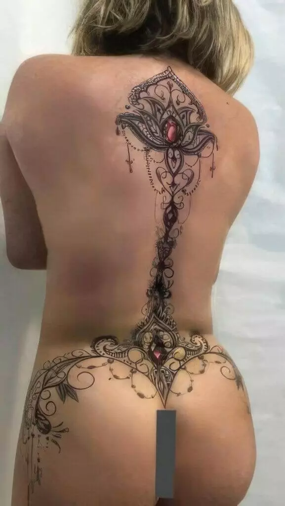 A person with their back turned, displaying an intricate back tattoo design featuring ornamental patterns and red gem-like elements stretching from the upper back to the lower back.