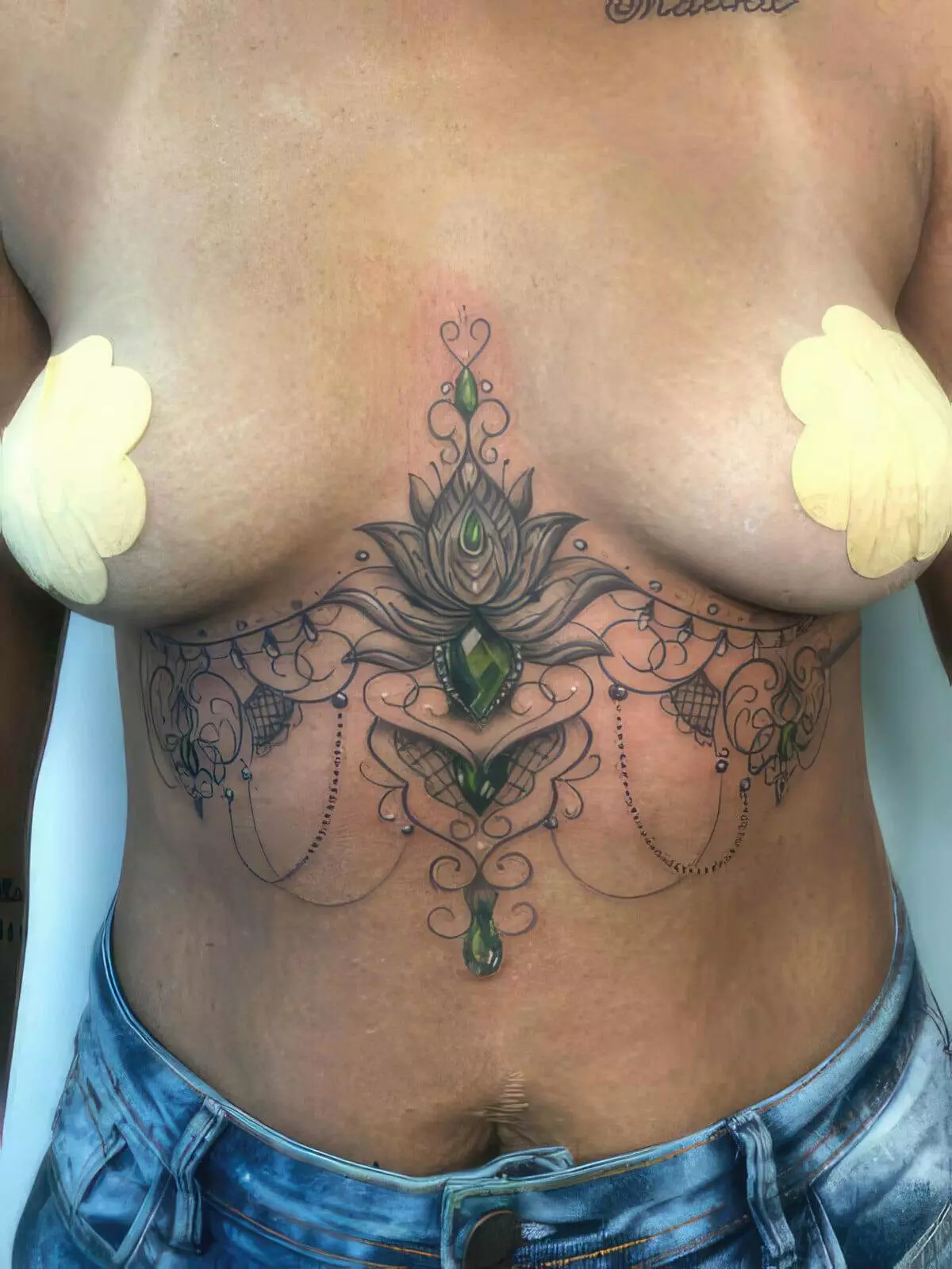 A person with a detailed chest and stomach tattoo featuring intricate designs and green gemstones. The individual is wearing light blue jeans and has adhesive covers on their breasts.