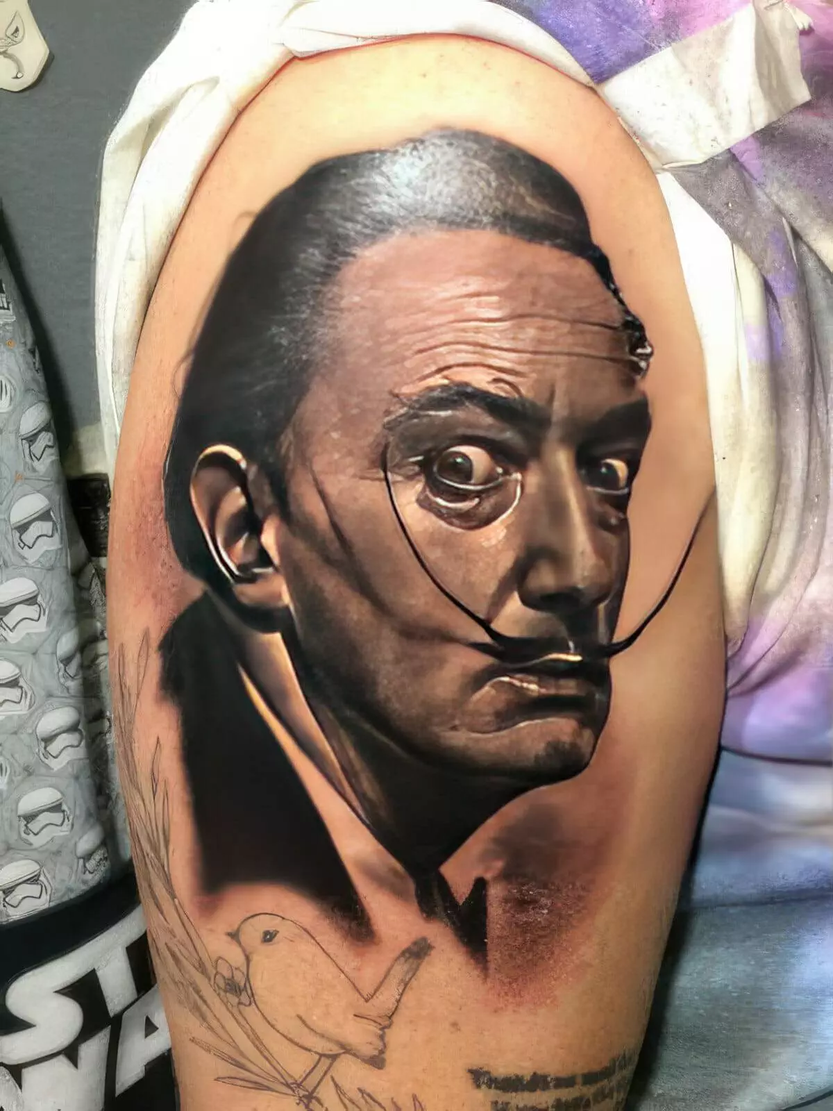 A detailed tattoo of a man's face with a distinctive mustache is shown on an arm, featuring precise shading and a partial sketch of a bird below.