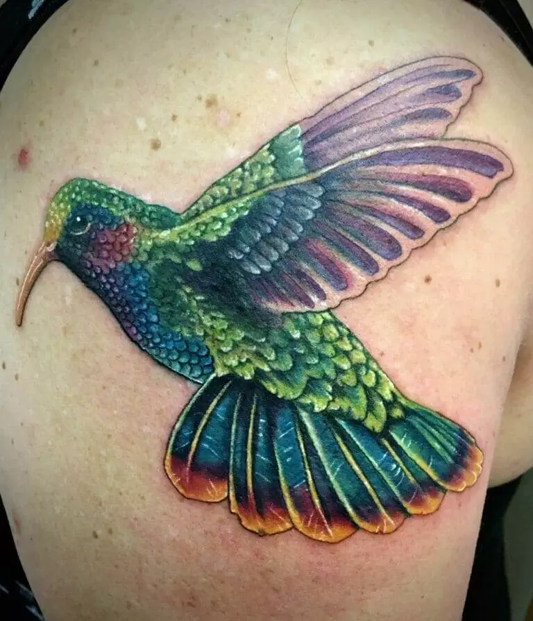 A colorful tattoo of a hummingbird in flight, displayed on a person's upper arm. The bird features green, blue, purple, and yellow hues.