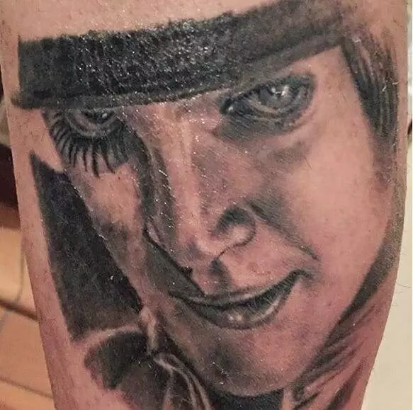 A detailed black and gray tattoo portrays a person's face wearing a hat, with an intense gaze and subtle smile.