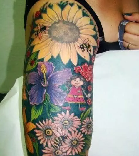 A colorful tattoo on an upper arm featuring various flowers, bees, and a cartoon-like girl in a dress.