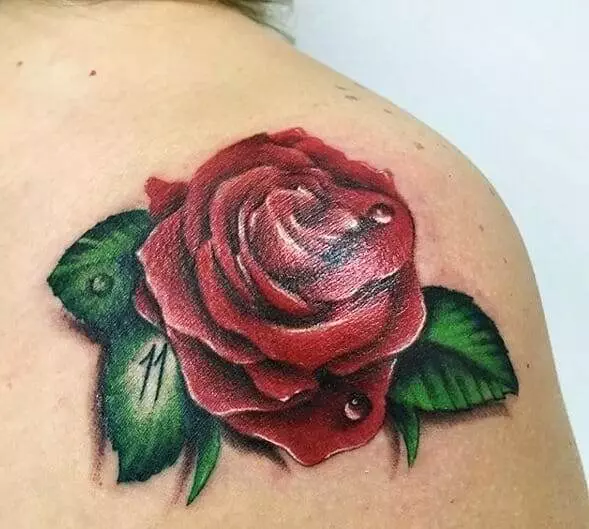 A realistic tattoo of a red rose with water droplets and green leaves on someone's shoulder.