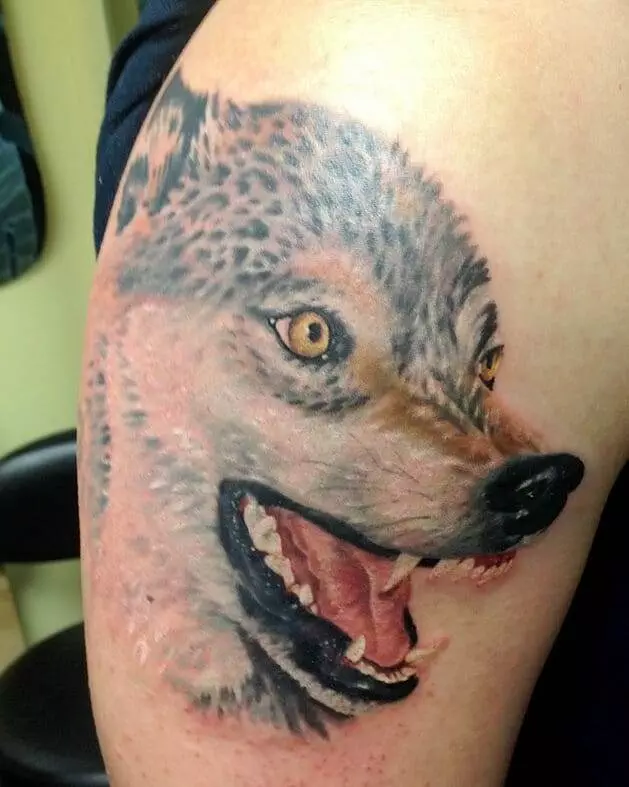 Detailed tattoo of a wolf's head with yellow eyes and an open mouth, located on a person's upper arm.