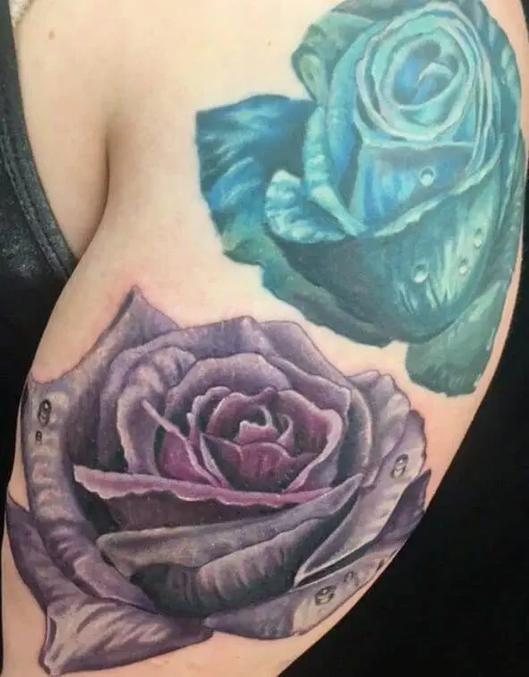 Close-up of an arm with two realistic rose tattoos, one purple and one blue, with water droplets on the petals.