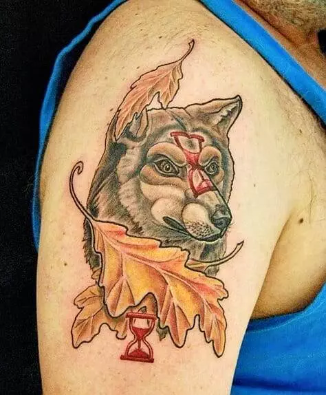 A tattoo of a wolf head with two red hourglasses and autumn leaves is displayed on the upper arm of a person wearing a blue tank top.