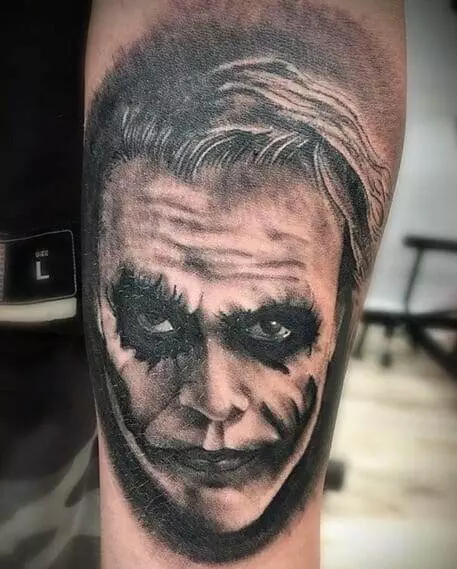 A detailed tattoo of a face with dark eye makeup and a sinister smile on someone's arm.