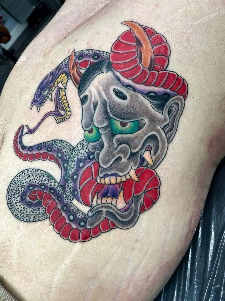 A detailed tattoo featuring a Hannya mask, with vibrant green eyes, interwoven with a coiled red and purple snake, and surrounded by additional snake elements on pale skin.