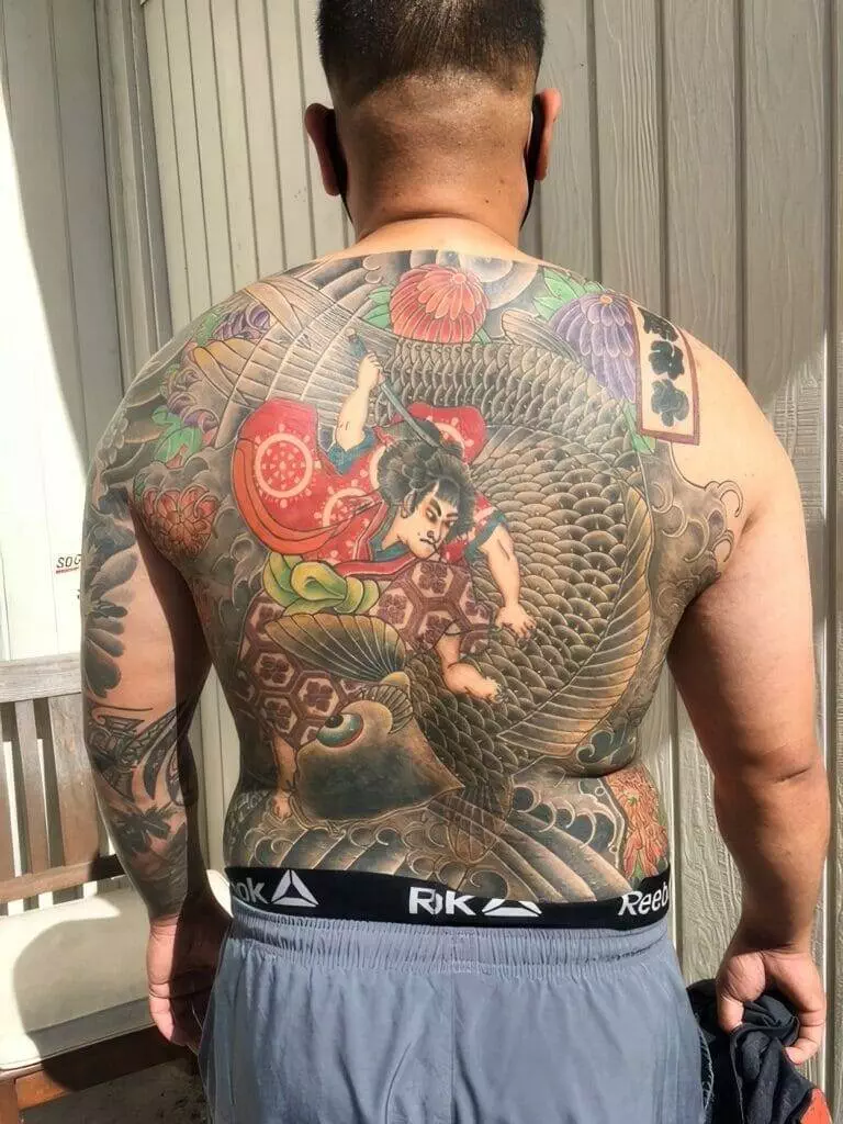 A person with an intricate tattoo covering their back features a colorful traditional Japanese design, including a warrior and dragon elements. The person is shirtless, wearing grey Reebok shorts.