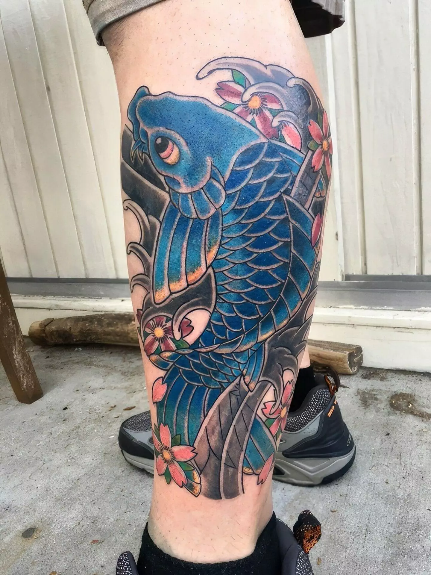 A colorful tattoo of a koi fish with flowers on a person's calf. The design features vibrant blues, pinks, and oranges against a wooden background.