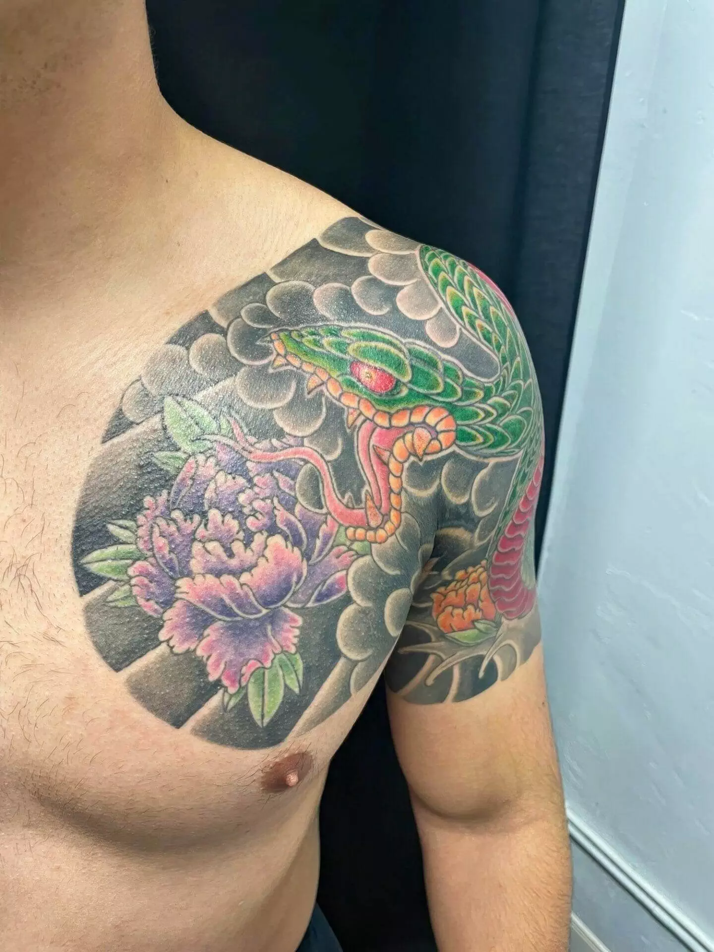 A person's shoulder and chest feature a vibrant tattoo of a snake wrapped around a pink and purple flower against a backdrop of dark clouds.