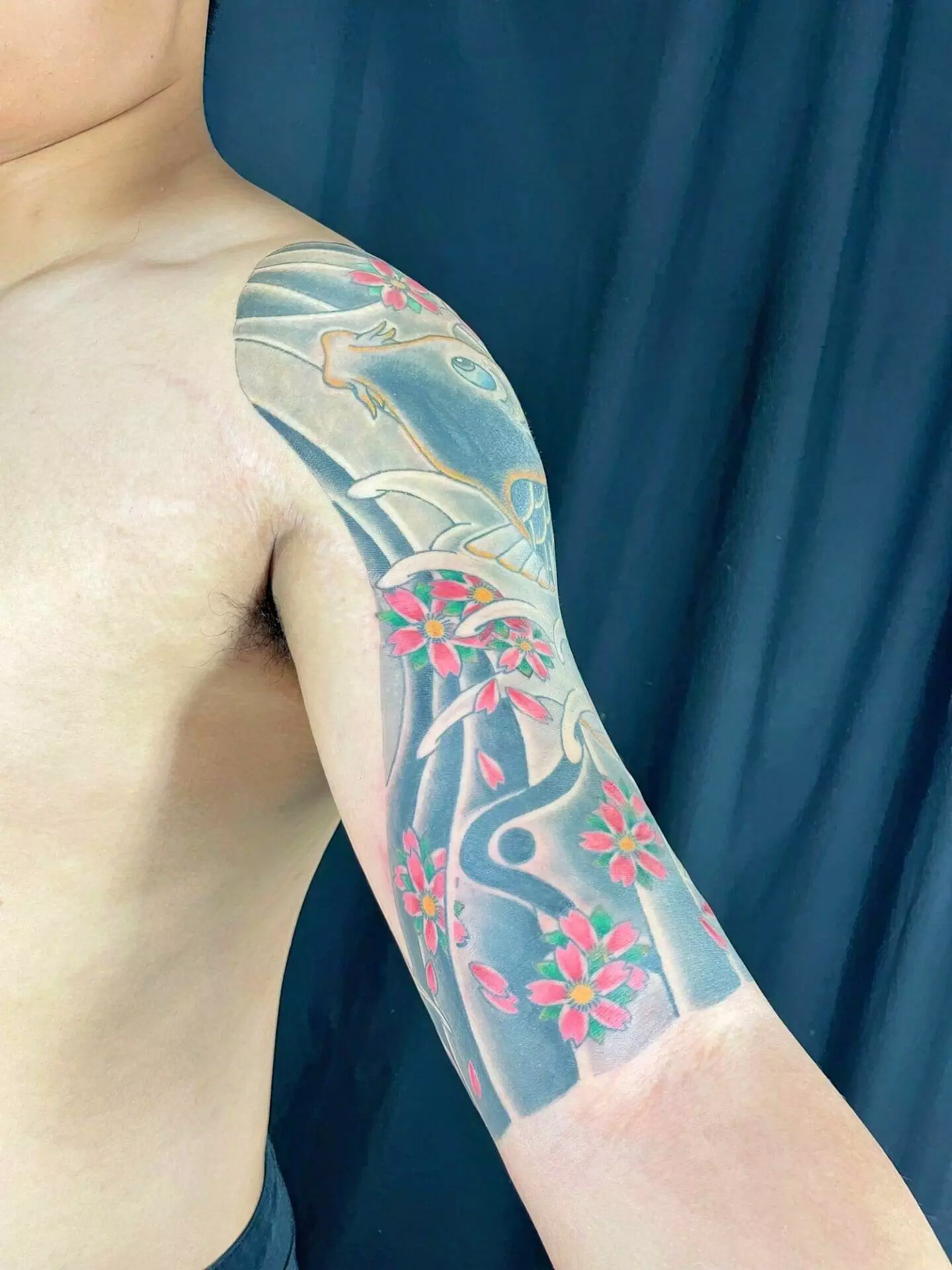 A person showcasing a colorful upper arm tattoo featuring pink flowers, leaves, and abstract designs against a dark background.