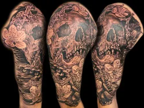 Tattoo featuring a detailed skull surrounded by flowers and various intricate designs, covering the upper arm and shoulder area.