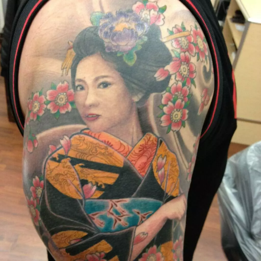 A detailed tattoo on an arm depicting a geisha with a floral background, wearing a traditional kimono with intricate patterns and vibrant colors.