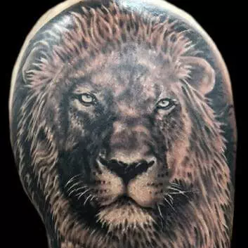 Realistic lion head tattoo with detailed fur and piercing eyes on a person's upper arm and shoulder.