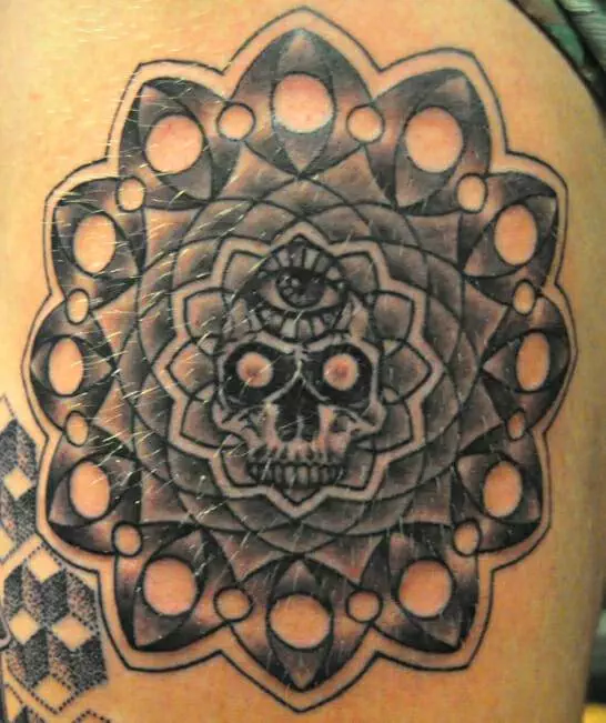 Black ink tattoo of a skull with an eye on its forehead, surrounded by geometric patterns and circular shapes on a person's skin.